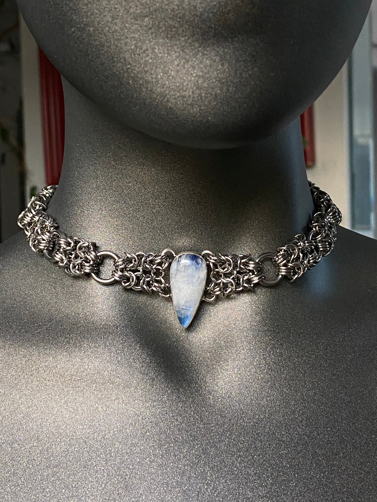 A silver-toned chainmail-style choker necklace with a central teardrop-shaped blue and white stone pendant, displayed on a matte gray mannequin bust.